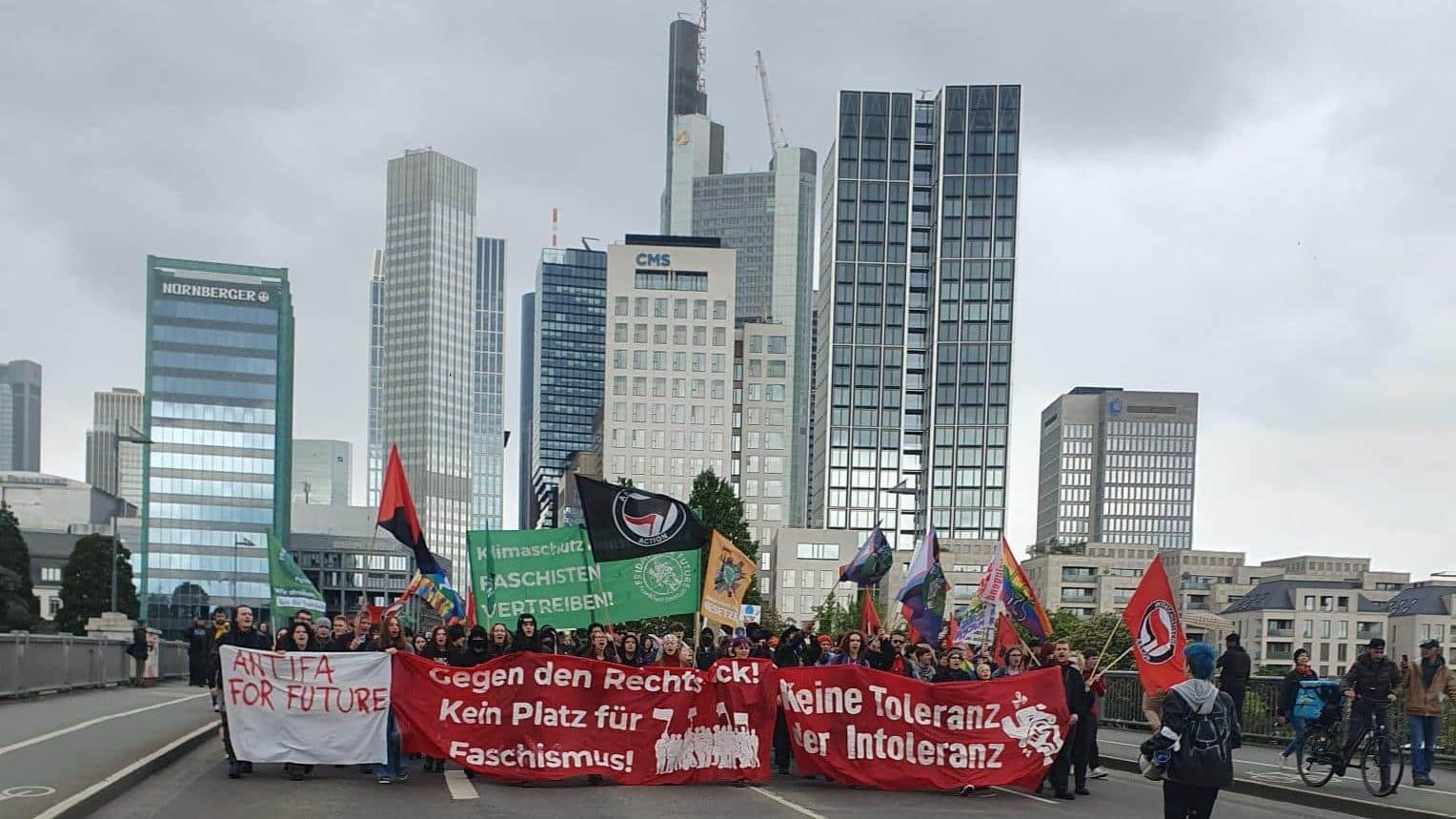 Hintergrund: Skyline von Frankfurt. Vordergrund: Demonstrierende mit Banner, auf dem steht: "Gegen den Rechtsruck! Kein Platz für Faschismus! Keine Toleranz der Intoleranz!"