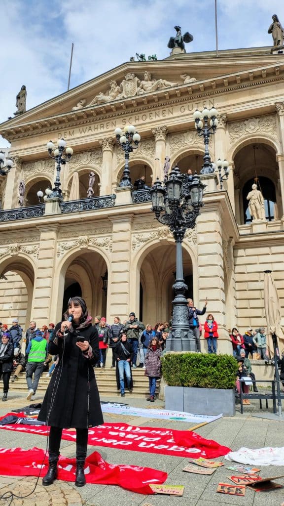 Das Foto zeigt Kerstin vom KoalaKollektiv, die mit einem Mikrofon vor der Alten Oper in Frankfurt steht. Auf dem Gebäude steht: "Dem Wahren Schönen Guten". Hinter Kerstin stehen Teilnehmende der Demo "zusammen gegen rechts".